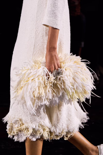 Zbliżenie na detal akcesorium na wybiegu: modelka trzyma puszystą, jasną kopertówkę wykonaną z miękkich piór w odcieniach bieli i kremu. Torebka kontrastuje z prostą, tweedową sukienką o surowym wykończeniu dołu, tworząc zestawienie delikatności i struktury w duchu haute couture.