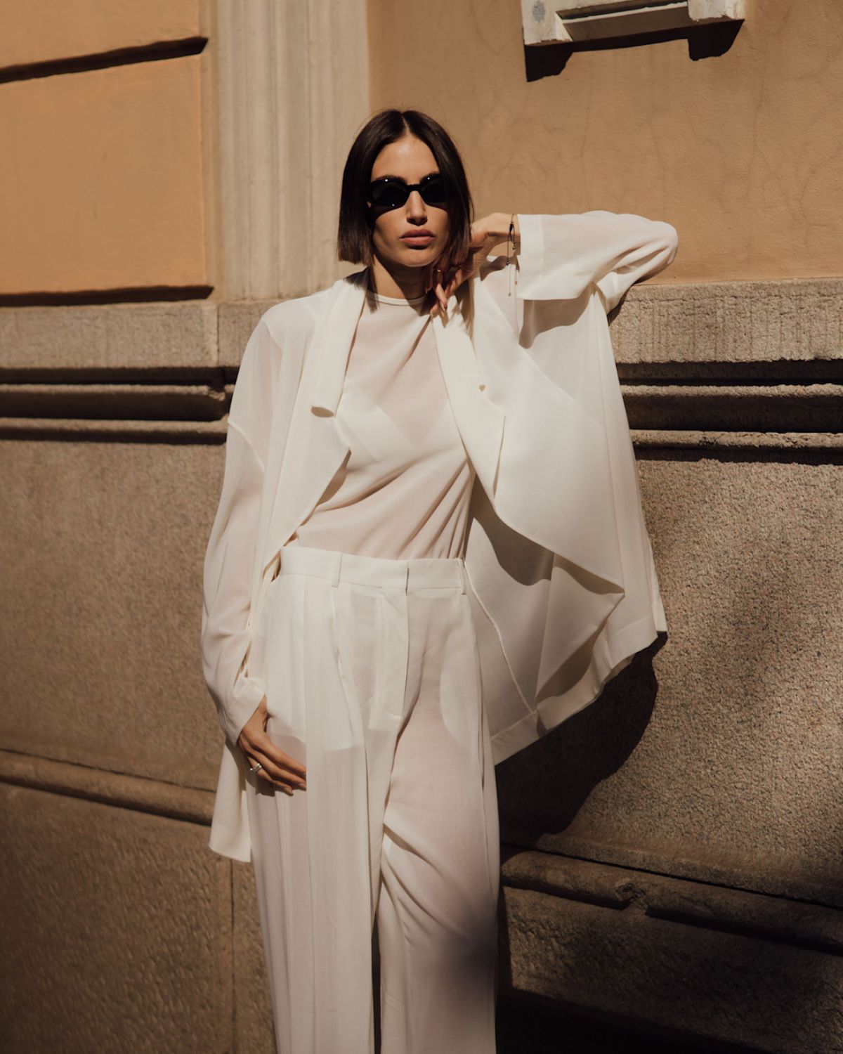 Kobieta w eleganckim, białym total looku składającym się z luźnych spodni palazzo i transparentnej koszuli, prezentująca, jak zbudować kapsułową garderobę opartą na ponadczasowych krojach i kolorach.
