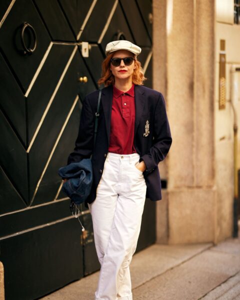 Kobieta w stylizacji nawiązującej do estetyki preppy i vintage, składającej się z granatowej marynarki z herbem, bordowej koszulki polo i białych spodni z wysokim stanem, uzupełnionej o stylowy kaszkiet oraz biało-czarne mokasyny – przykład na to, jak zbudować kapsułową garderobę z charakterem.