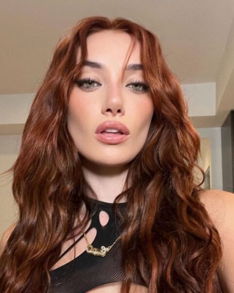 Portret kobiety o oliwkowej cerze, prezentującej głęboki, miedziany odcień kasztana, który idealnie wpisuje się w kolory włosów 2026 roku. Długie, pofalowane pasma z przedziałkiem na środku okalają twarz z wyrazistym makijażem typu „cat eye” i ustami w kolorze nude. Stylizację dopełnia czarny top z modnymi wycięciami oraz złoty naszyjnik, tworząc zmysłowy i nowoczesny wizerunek.