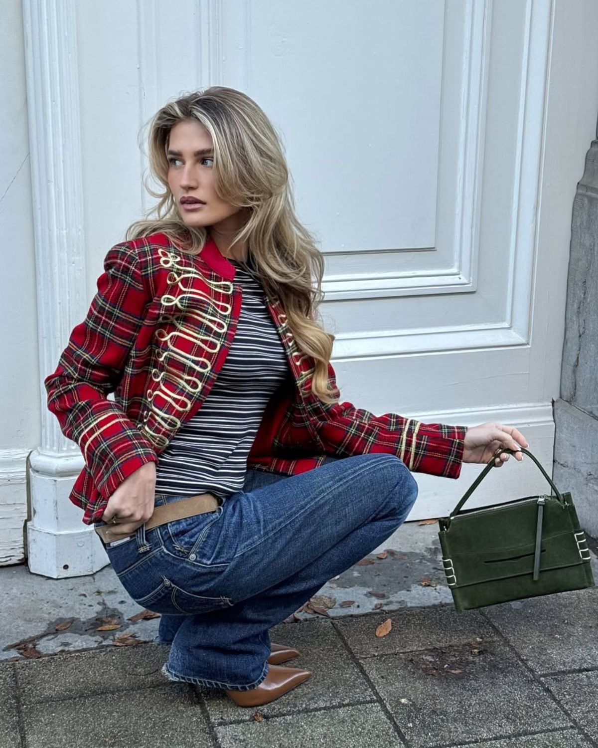 Kobieta w inspirującej stylizacji łączącej modne wzory ubrań 2026: czerwoną marynarkę w kratę o fasonie militarnym ze złotymi zdobieniami oraz czarno-biały prążkowany longsleeve. Całość dopełniają klasyczne jeansy, brązowe szpilki w szpic i elegancka, zamszowa torebka w kolorze butelkowej zieleni na tle białych, stylowych drzwi.
