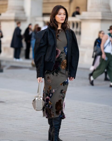 Kobieta podczas Paris Fashion Week prezentuje stylizację typu street style, na którą składa się długa, ciemnooliwkowa sukienka w nasycone, wielobarwne kwiatowe wzory oraz czarna kurtka bomberka o architektonicznej formie z wyraźnie zaznaczonymi ramionami. Całość uzupełniają wysokie, czarne skórzane kozaki na słupku oraz elegancka, szaro-beżowa torebka o geometrycznym kształcie, trzymana w dłoni.