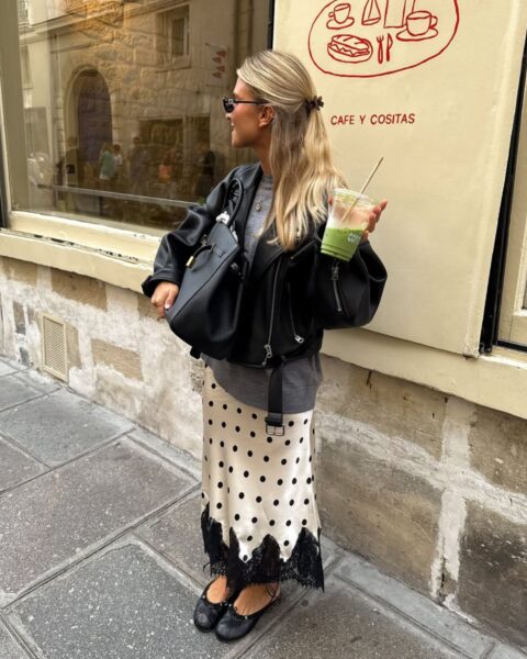Młoda kobieta w paryskiej stylizacji street style, stojąca przed kawiarnią z kubkiem matcha latte. Ma na sobie satynową spódnicę midi w kolorze kości słoniowej w czarne grochy, wykończoną szeroką, czarną koronką. Zestaw uzupełnia szary t-shirt, oversize’owa czarna skórzana kurtka ramoneska oraz czarne baleriny z wiązaniem. W dłoni trzyma dużą, czarną skórzaną torbę typu tote.
