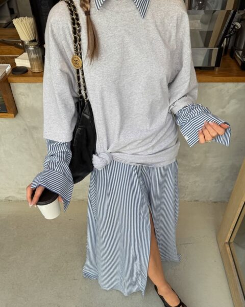 Zbliżenie na kreatywną, warstwową stylizację w stylu paryskim. Kobieta ma na sobie błękitną sukienkę koszulową w pionowe białe prążki, na którą założony jest szary, bawełniany t-shirt oversize fantazyjnie związany na boku w supeł. Spod krótkich rękawów koszulki wystają szerokie mankiety sukienki. Całość uzupełnia czarna torebka na złotym łańcuszku z ozdobnym medalionem oraz modne, ażurowe balerinki typu "mesh".