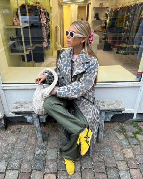 Kobieta siedząca na drewnianej ławce przed butikiem, prezentująca odważną, miejską stylizację. Ma na sobie długi płaszcz typu trencz w wyrazisty wzór panterki w odcieniach szarości i brązu, luźne spodnie cargo w kolorze oliwkowym oraz jaskrawożółte sportowe sneakersy. Całość uzupełniają białe okulary przeciwsłoneczne w grubej oprawie, różowa ozdobna spinka-kwiat we włosach oraz duża, pleciona torebka typu „cloud bag” w kolorze kremowym.