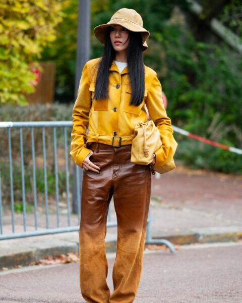 Inspirująca stylizacja street style w odcieniach miodu i brązu z Paris Fashion Week. Kobieta ma na sobie żółtą zamszową kurtkę z paskiem, połączoną z szerokimi, dwukolorowymi spodniami o efekcie ombre (ciemnobrązowa lakierowana góra przechodząca w jasny zamsz na dole). Całość uzupełnia beżowy kapelusz typu bucket hat, torebka typu „pouch” w kolorze miodowym oraz brązowe botki w szpic.