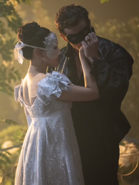 Romantyczna scena balowa w plenerze: młoda kobieta ubrana w jasną, połyskującą suknię z delikatnymi rękawkami i wiązaniem na plecach tańczy z mężczyzną w ciemnym, historyzującym stroju. Oboje mają na twarzach maski balowe, a ich sylwetki otula miękkie, złotawe światło przenikające przez liście drzew, tworząc nastrojową, niemal baśniową atmosferę inspirowaną estetyką XVIII wieku.