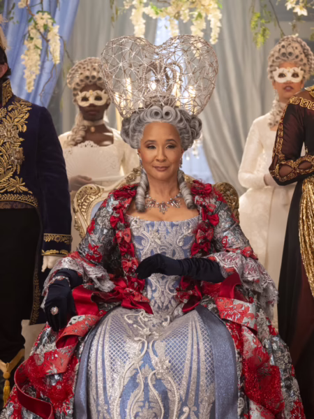 Centralna postać siedząca na ozdobnym tronie podczas wystawnego balu dworskiego: dojrzała kobieta w bogato haftowanej sukni w odcieniach błękitu, srebra i czerwieni, z dekoracyjnymi aplikacjami i długimi rękawami. Jej stylizację dopełnia imponująca, rzeźbiarska peruka w stylu XVIII wieku oraz biżuteria. W tle stoją elegancko ubrani goście w maskach, tworząc symetryczną, ceremonialną kompozycję.