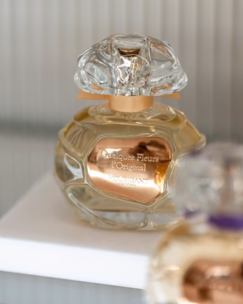 uksusowy, rzeźbiony flakon perfum Quelques Fleurs l’Original marki Houbigant Paris ze złotą etykietą i dekoracyjnym korkiem przypominającym kwiat, prezentowany na minimalistycznym, białym podestacie.