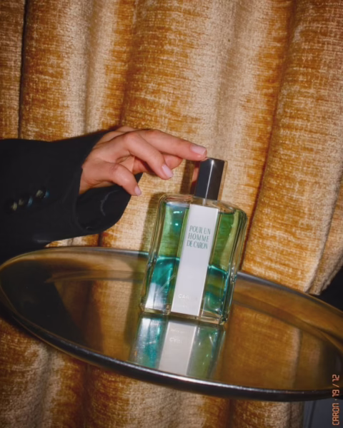 Emanujący elegancją flakon klasycznego zapachu Pour Un Homme de Caron, umieszczony na srebrnej tacy na tle miodowej, aksamitnej zasłony. Zdjęcie w stylu retro, na którym widać dłoń w czarnej marynarce, ilustruje trend, w którym wracają stare marki perfumeryjne.