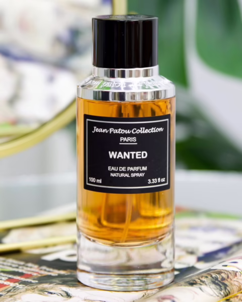 Elegancki flakon wody perfumowanej Jean Patou Collection Paris „Wanted” o pojemności 100 ml, wypełniony bursztynowym płynem. Minimalistyczna butelka z czarną etykietą i srebrnymi detalami stoi na kolorowych magazynach modowych, w tle widoczne są rozmyte zielone liście roślin, co podkreśla luksusowy charakter produktu.