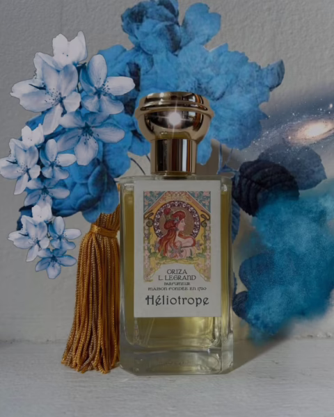 Artystyczne ujęcie flakonu perfum Héliotrope marki Oriza L. Legrand z ozdobną etykietą w stylu secesyjnym, przedstawiającą kobietę wśród kwiatów. Butelka z eleganckim złotym korkiem i chwostem stoi na tle błękitnych kwiatów i mistycznej, niebieskiej mgły, co doskonale obrazuje zjawisko, w którym wracają stare marki perfumeryjne do świata współczesnego luksusu.