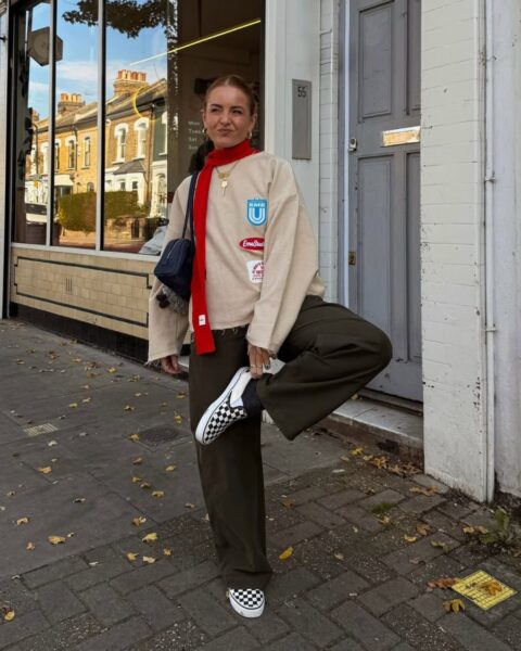 Kobieta w jesiennym outficie stoi na chodniku przed kamienicą, pozując na jednej nodze; ma na sobie beżową bluzę z naszywkami, szerokie spodnie w kolorze khaki, czerwony wąski szal oraz czarno-białe wsuwane tenisówki w szachownicę.