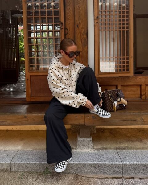 Kobieta w okularach przeciwsłonecznych siedzi na drewnianym tarasie tradycyjnego budynku, poprawiając buty; ma na sobie beżową bluzę z kapturem w czarne grochy, szerokie czarne spodnie oraz wsuwane tenisówki w charakterystyczną szachownicę; obok stoi luksusowa torebka w brązową kratę.
