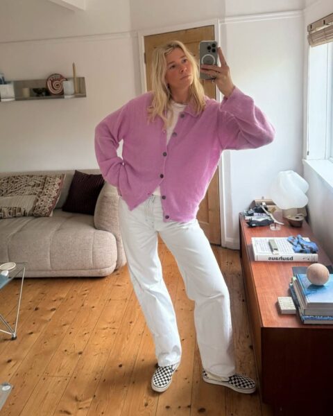 Zdjęcie typu „selfie w lustrze” przedstawiające kobietę w jasnym, minimalistycznym wnętrzu; ma na sobie rozpięty liliowy kardigan, biały t-shirt, jasne luźne jeansy oraz wsuwane tenisówki w czarno-białą szachownicę; w tle widoczna nowoczesna beżowa sofa z wzorzystymi poduszkami oraz drewniana komoda z designerską lampą i książkami.