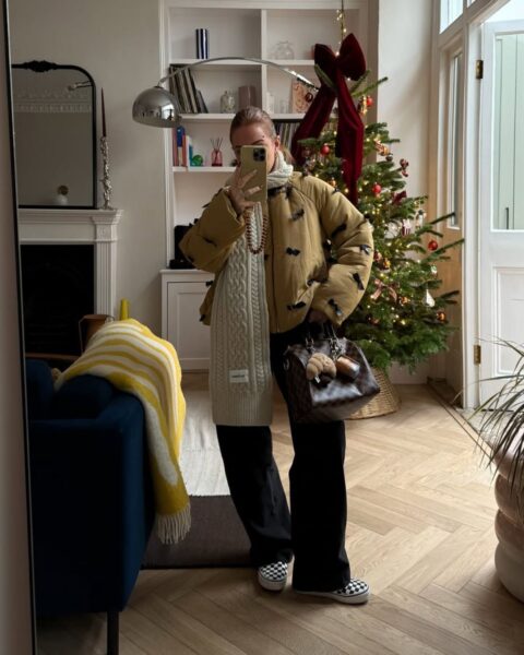 Kobieta wykonuje selfie w lustrze w eleganckim salonie z choinką; ma na sobie musztardową kurtkę puchową z czarnymi kokardkami, gruby kremowy szal o wyraźnym splocie, ciemne spodnie oraz czarno-białe wsuwane tenisówki wróciły do mody jako baza zimowych stylizacji; w tle widać stylowy kominek, biały regał z książkami i designerskie dodatki.