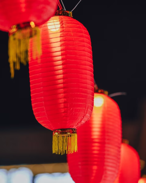 Rząd podłużnych, czerwonych lampionów z żółtymi frędzlami, delikatnie rozświetlonych od środka. Pierwszy plan jest ostry, ukazując teksturę materiału, podczas gdy kolejne lampiony tworzą nastrojowy efekt bokeh na ciemnym tle, budując klimat wieczornego świętowania.