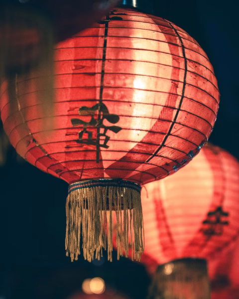Nastrojowe zbliżenie na tradycyjny, czerwony lampion z czarnymi znakami chińskiej kaligrafii i złotymi frędzlami. Ciepłe światło przenikające przez papierową strukturę tworzy intymny klimat i głębię na ciemnym tle, idealnie oddając estetykę wieczornych celebracji.