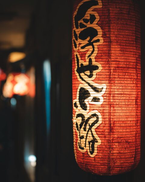 Zbliżenie na pionowy, cylindryczny lampion w kolorze nasyconej czerwieni, ozdobiony dużą, czarną kaligrafią na białym tle. Lampion jest rozświetlony od wewnątrz, a ostrość skupiona na fakturze papieru i znakach, podczas gdy tło pozostaje ciemne i nastrojowo rozmyte.