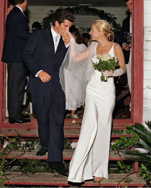 Kennedy ślub: John F. Kennedy Jr. i Carolyn Bessette-Kennedy wychodzą z drewnianej kaplicy. Carolyn ma na sobie jedwabną suknię ślubną Narciso Rodriguez, długie tiulowe rękawiczki i trzyma bukiet z konwalii, podczas gdy John czule całuje ją w dłoń.