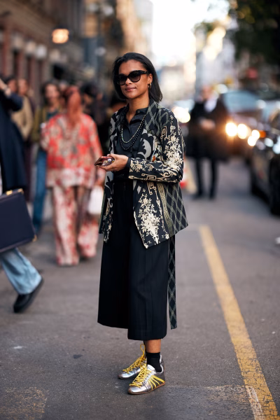 Metaliczne sneakersy ze złotymi detalami przełamują klasyczny, czarny look z marynarką i szerokimi spodniami. To przykład, jak modne sneakersy na wiosnę 2026 w wersji srebrnej lub złotej dodają stylizacji futurystycznego charakteru.