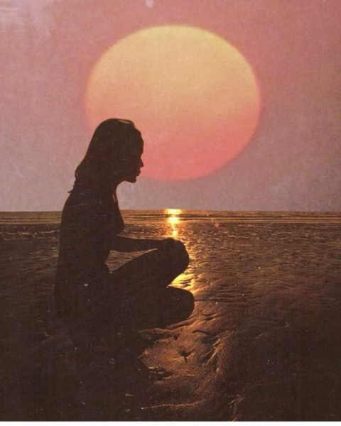 Artystyczne ujęcie ciemnej sylwetki kobiety kucającej na mokrym piasku plaży w świetle zachodzącego słońca. Nad horyzontem góruje ogromna, bladoróżowa tarcza słońca, której blask odbija się złotą smugą na powierzchni wody i wilgotnym brzegu. Całość utrzymana w ciepłej, nastrojowej estetyce retro z delikatną teksturą ziarna.