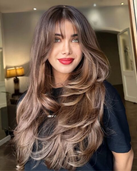 Portret młodej kobiety z długimi, gęstymi, cieniowanymi włosami w kolorze jasnego brązu z karmelowymi refleksami. Włosy ułożone są w puszyste fale, a na czole widać delikatną grzywkę typu "curtain bangs". Kobieta ma zielone oczy i jaskrawoczerwoną szminkę na ustach. Nosi czarną koszulkę. Tło jest lekko rozmyte, widać wnętrze salonu fryzjerskiego z lampą w kształcie dyni i białymi drzwiami.