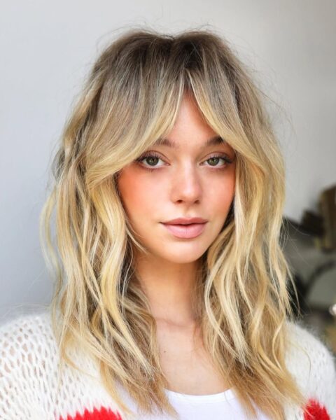 Portret młodej kobiety o jasnych, miodowych blond włosach z ciemniejszym odrostem, ułożonych w puszystą fryzurę typu "shaggy hair" z wyraźną grzywką "curtain bangs". Włosy mają naturalną, falowaną strukturę i dużą objętość. Kobieta ma brzoskwiniowy makijaż policzków i ust, a ubrana jest w biały, ażurowy sweter z czerwonymi akcentami. Tło jest jasne i minimalistyczne.