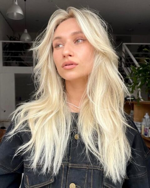 Portret kobiety o bardzo jasnych, chłodnych blond włosach, sięgających za ramiona. Fryzura charakteryzuje się mocnym cieniowaniem w stylu „butterfly cut” z długą, rozchodzącą się na boki grzywką typu „curtain bangs”, która nadaje włosom dużą objętość i lekkość. Kobieta ma delikatny makijaż w odcieniach nude i patrzy w bok. Ubrana jest w czarną, jeansową kurtkę z widocznymi przeszyciami oraz subtelny naszyjnik. Tło przedstawia jasne, nowoczesne wnętrze z antresolą i roślinami.