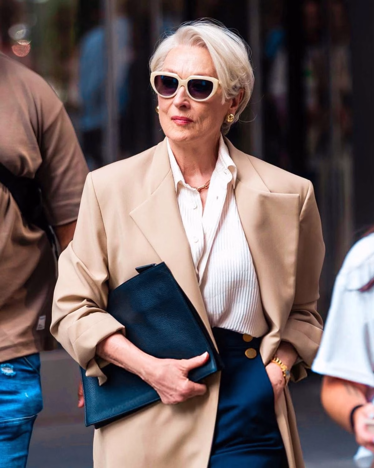 Elegancka starsza kobieta w miejskiej scenerii, ubrana w beżowy płaszcz i białą koszulę, z krótkimi siwymi włosami i dużymi okularami przeciwsłonecznymi, trzyma czarną kopertówkę — styl inspirowany trendy z filmu „Diabeł ubiera się u Prady 2”.