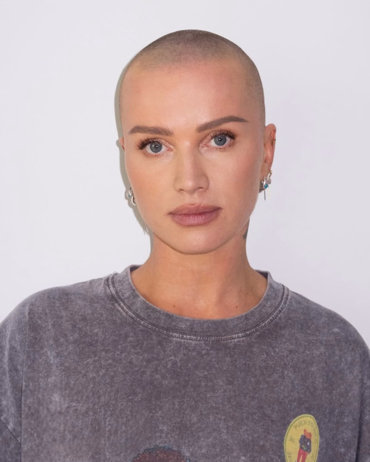 Portret kobiety ogolonej na łyso, patrzącej prosto w obiektyw, w szarej koszulce i kolczykach na jasnym tle – wszystkie gwiazdy, które ogoliły głowę.