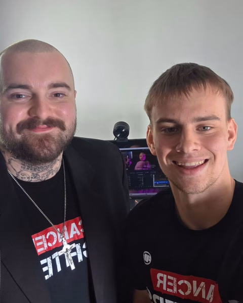 Bedoes i Łatwogang uśmiechają się do zdjęcia w domowym studiu, stojąc przed monitorem komputera, ubrani w czarne koszulki z nadrukiem.