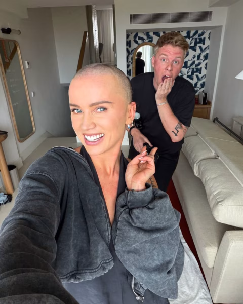 Influencerka Maffashion robi uśmiechnięte selfie w hotelowym pokoju po ogoleniu głowy, a stojący za nią mężczyzna reaguje zaskoczeniem.