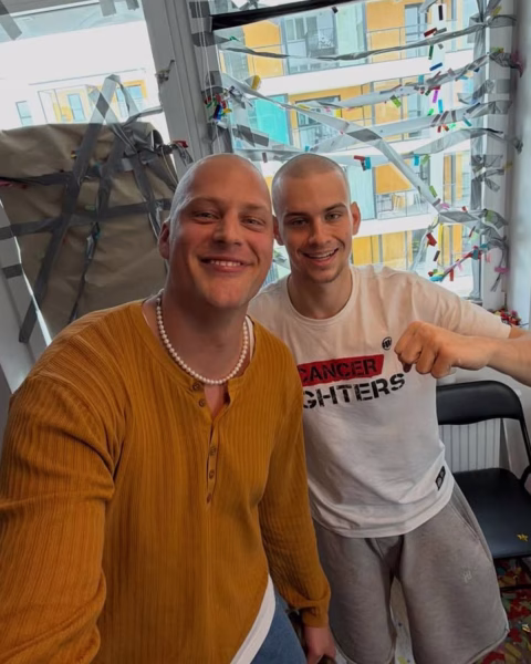 Influencer Jakob Kosel i towarzyszący mu Łatwogang pozują do uśmiechniętego selfie w mieszkaniu, obaj z ogolonymi głowami, na tle okna udekorowanego taśmami i konfetti.