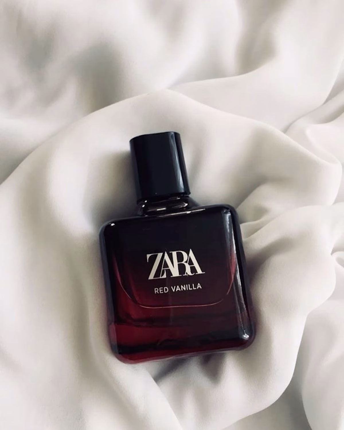 Perfumy Zara odpowiedniki. Zdjęcie Zara Red Vanilla – flakon perfum w ciemnym, czerwono-czarnym odcieniu ułożony na miękkiej, jasnej tkaninie. Elegancki design podkreśla ciepły, słodki i otulający charakter zapachu.