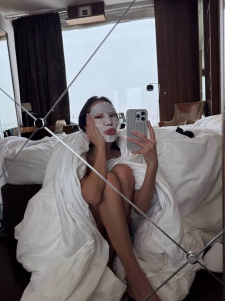 Kobieta w maseczce w płachcie robiąca selfie w łóżku, co pokazuje moment intensywnego nawilżenia typowy dla koreańskich protokołów pielęgnacyjnych. Tego typu maska wspiera regenerację skóry i odbudowę bariery hydrolipidowej, szczególnie w protokole nawilżającym lub kojącym. To przykład pielęgnacji dopasowanej do potrzeb skóry – prosty, ale skuteczny etap rutyny K-beauty.