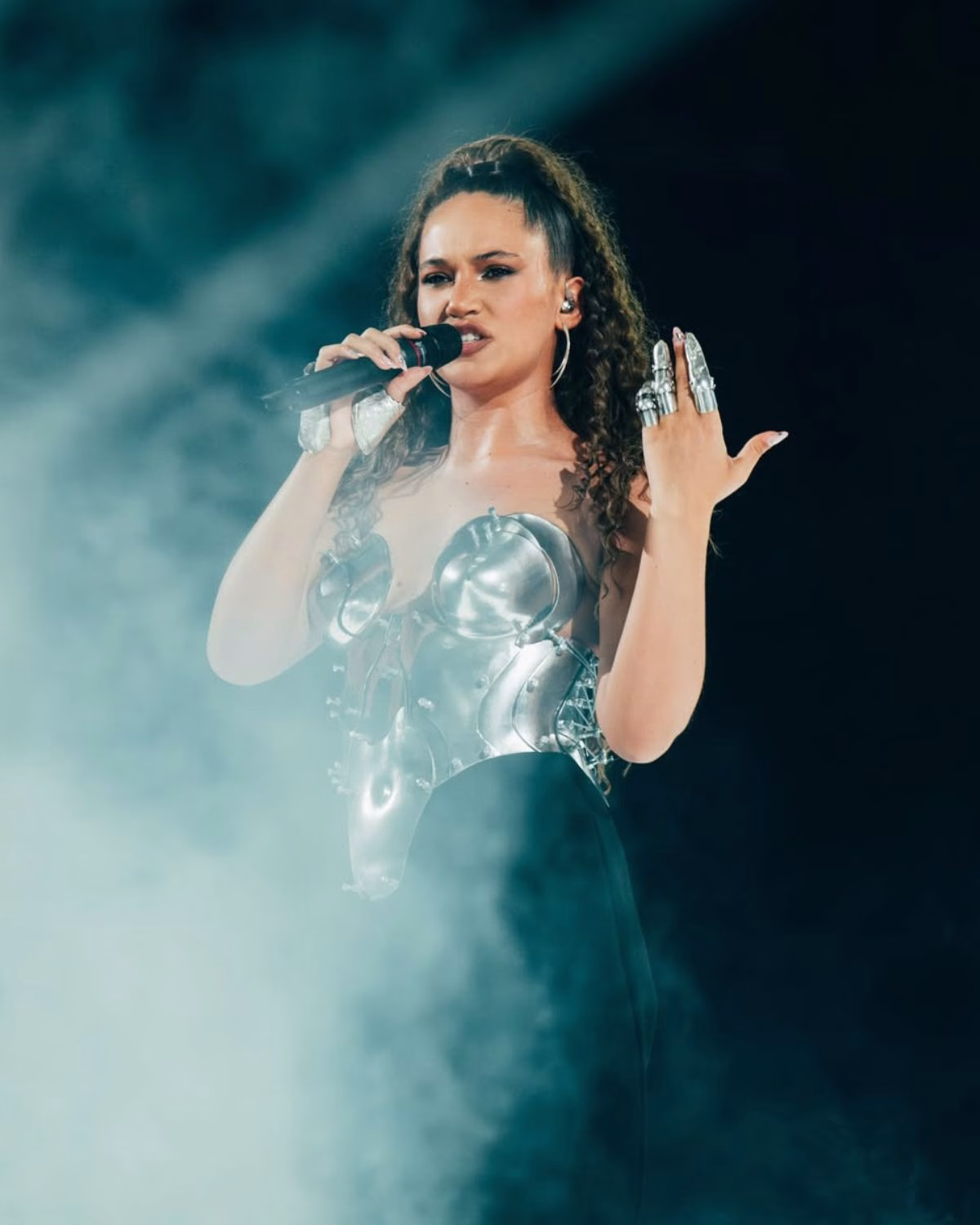 Alicja Szemplińska stylizacja Eurowizja 2026 w trakcie występu scenicznego. Wokalistka w srebrnym gorsecie i czarnych spodniach śpiewa do mikrofonu, a dym otacza scenę. Na jej dłoni widać ozdobne, metalowe nakładki na palce.