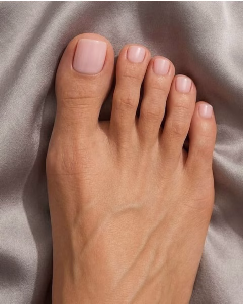 Zbliżenie na zadbaną stopę z eleganckim, minimalistycznym pedicure, opierającą się na szarej, gładkiej tkaninie. Paznokcie u stóp mają kwadratowy kształt z delikatnie zaokrąglonymi rogami i są pomalowane na jasny, półtransparentny, mlecznoróżowy kolor o subtelnym połysku. Skóra stopy ma naturalny odcień, a palce są prosto ułożone, prezentując estetyczne wykonanie stylizacji paznokci.