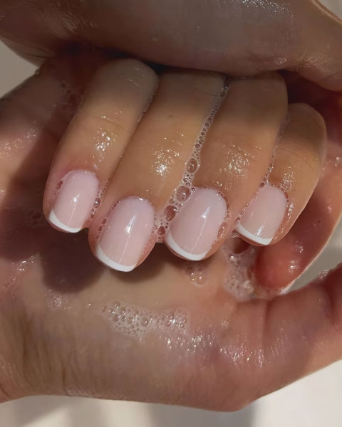 Zbliżenie na dłoń ze świeżym manicure francuskim o krótkim, naturalnym kształcie, z mlecznoróżową bazą i białą końcówką. Palce są ułożone blisko siebie i zanurzone w dłoni pokrytej białą pianą mydlaną z małymi bąbelkami, która otacza płytki paznokci. Zdjęcie ma ciepłe, naturalne oświetlenie, a tło jest rozmyte.