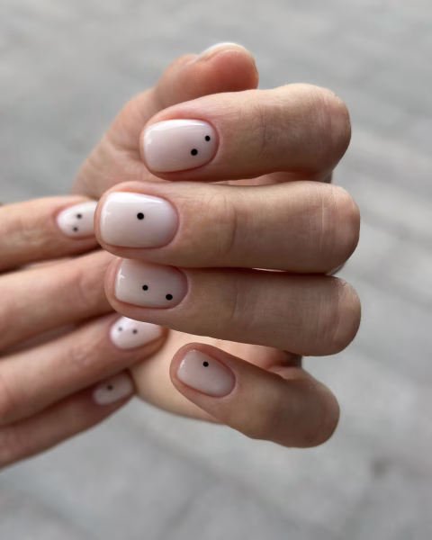 Zbliżenie na dłonie z minimalistycznym manicure na krótkich paznokciach. Paznokcie są pomalowane błyszczącym, mlecznobiałym lakierem hybrydowym. Każdy paznokieć ozdobiony jest jedną lub kilkoma małymi, czarnymi kropkami, umieszczonymi w różnych miejscach na płytce. Lewa dłoń spoczywa na prawej, a w tle widać rozmytą powierzchnię chodnika.