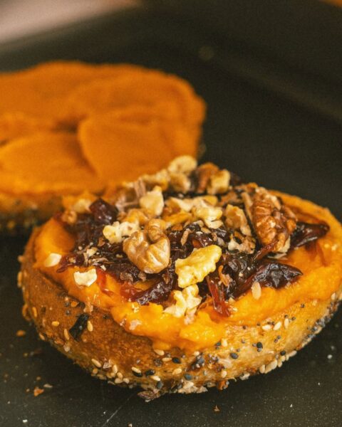 Bajgiel z posypką (makiem i sezamem) przekrojony na pół, z wierzchu obficie posmarowany pomarańczowym kremem dyniowym, a na nim widoczne orzechy włoskie i ciemna konfitura (prawdopodobnie żurawinowa). Zdjęcie przedstawia pożegnanie z jesiennym smakiem dyni przed wprowadzeniem zimowej oferty.