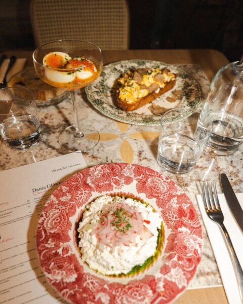 Zdjęcie śniadania na marmurowym stole: na pierwszym planie duży talerz z białą ricottą lub twarożkiem, udekorowany czerwoną cebulą i szczypiorkiem. Powyżej w kieliszku do koktajli jajka w koszulce z kawiorem i sosem, a obok na kwiecistym talerzu jajecznica na grzance z tartymi truflami. Obok leży menu, a na stole widoczne są szklanki z wodą.