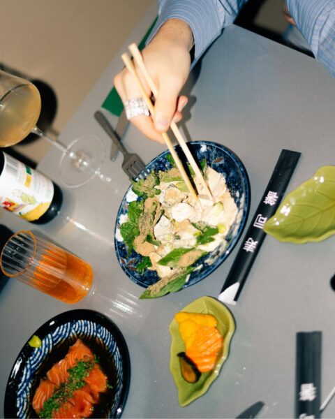 Zdjęcie z lotu ptaka przedstawiające rękę w koszuli w prążki, z dużym pierścieniem na palcu, trzymającą drewniane pałeczki nad miską z sałatką (prawdopodobnie z kurczakiem/tofu w kremowym sosie z miętą). Na stole widoczne są również: mały talerzyk z kawałkiem łososia, talerz z kawałkami łososia w sosie sojowym i szczypiorkiem, kieliszek z winem, butelka i czarny futerał na pałeczki leżący na zielonej podstawce w kształcie liścia. Scena kolacji/lunchu w azjatyckim stylu.