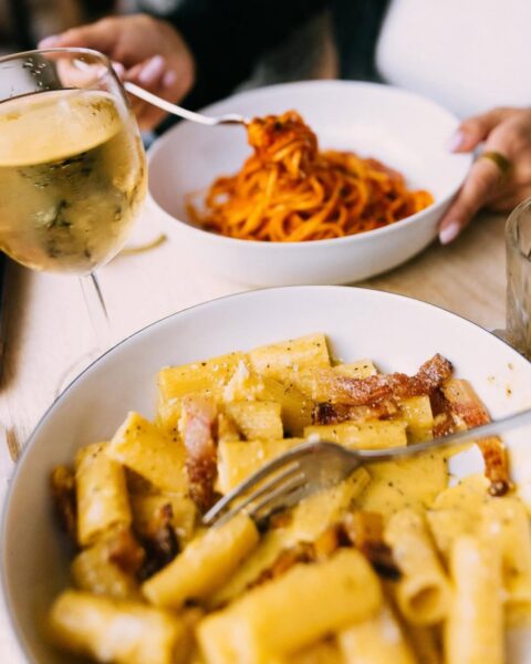 Dwa talerze z makaronem na jasnym stole: na pierwszym planie biała miska z makaronem typu rigatoni lub penne w kremowym, serowym sosie z kawałkami boczku. W misce widoczny widelec. W tle, częściowo zamazana, druga biała miska z długim, czerwonym makaronem (spaghetti lub linguine) w sosie pomidorowym, z widoczną dłonią z widelcem. Obok na stole stoi kieliszek z białym winem i szklanka.