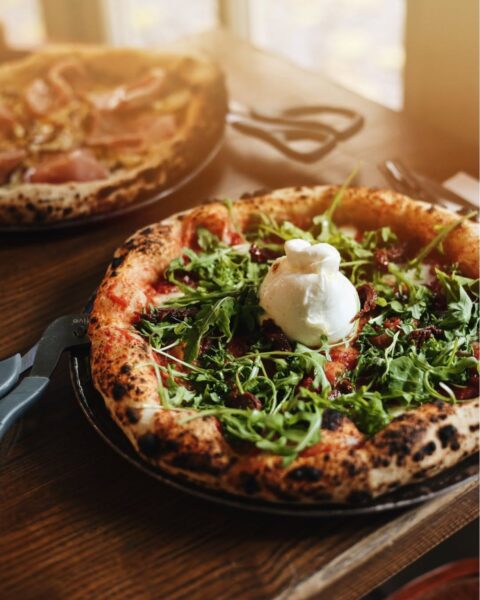 Pizza na cienkim cieście z mocno wypieczonymi, pęcherzykowatymi brzegami, na wierzchu posypana rukolą, suszonymi pomidorami i zwieńczona kulką białej burraty. Pizza leży na ciemnym talerzu na drewnianym stole w świetle wpadającym z okna. W tle, niewyraźna, widoczna jest druga pizza (prawdopodobnie z szynką).